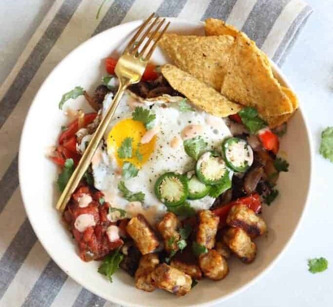 A wholesome Vegetarian Southwest Breakfast Bowl with a sunny-side-up egg, diced tomatoes, sliced jalapeños, and golden-brown roasted potatoes, garnished with fresh cilantro leaves and served with a side of crispy tortilla chips, presented on a white plate with a gold fork on a striped grey and white cloth. Vegetarian Southwest Breakfast Bowl