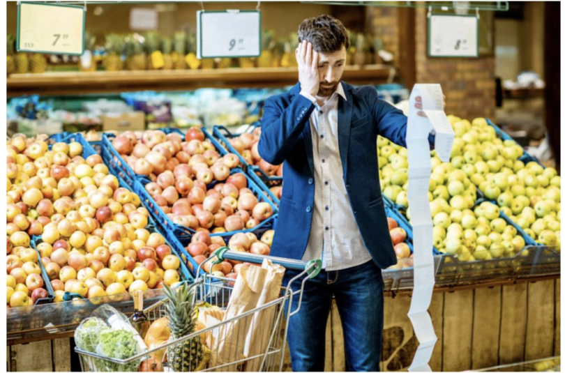 expensive grocery receipt Man in supermarket with shopping cart looking overwhelmed by a long grocery receipt in the fresh produce section.